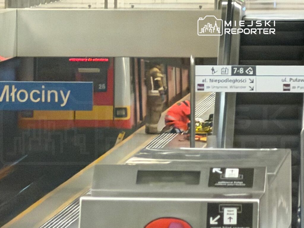 Pracownik w pomarańczowej odzieży roboczej dokonuje naprawy na stacji metra Młociny, obok pociągu.