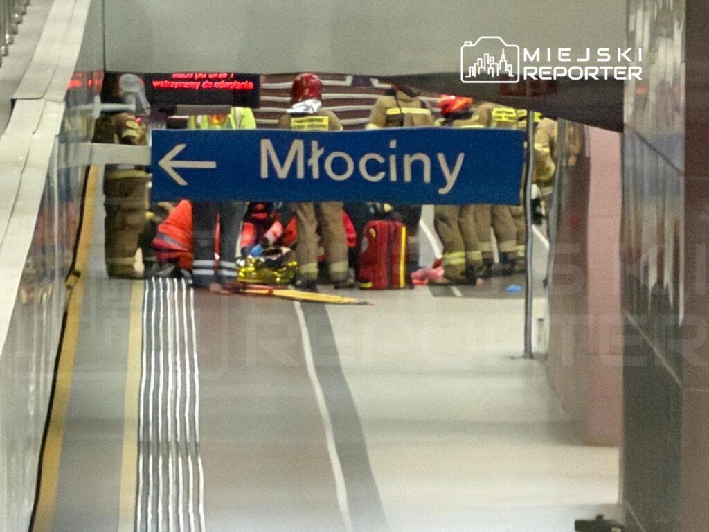 Grupa ratowników i strażaków udziela pomocy osobie na stacji metra, obok znaku kierunkowym "Młociny".
