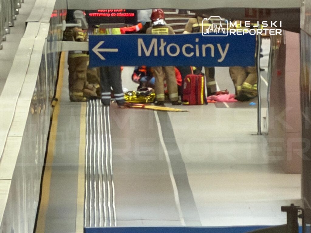 Grupa ratowników w mundurach udziela pomocy osobie leżącej na stacji metra, obok znaku kierunkowym "Młociny".