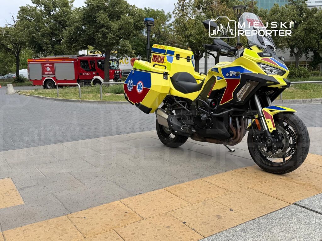 Motocykl ratunkowy w kolorze żółtym z czerwonymi i niebieskimi detalami stoi na chodniku, w tle widać wóz strażacki.