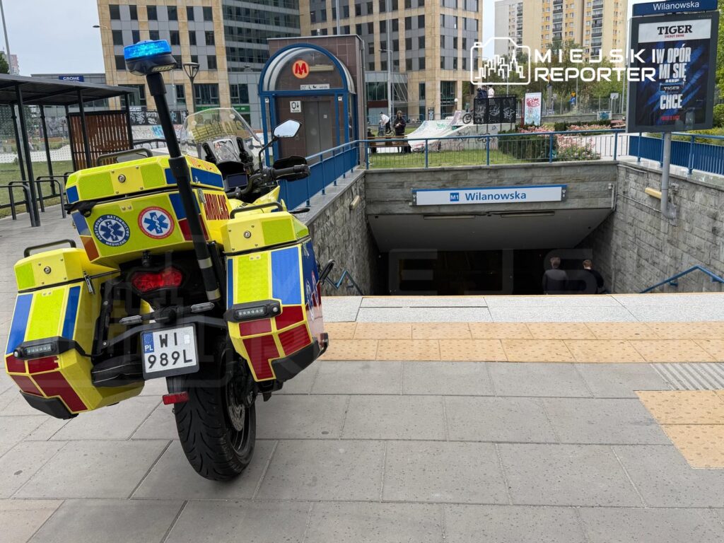 Motocykl służby ratunkowej z niebieskim światłem zaparkowany przy wejściu do stacji metra Wilanowska.