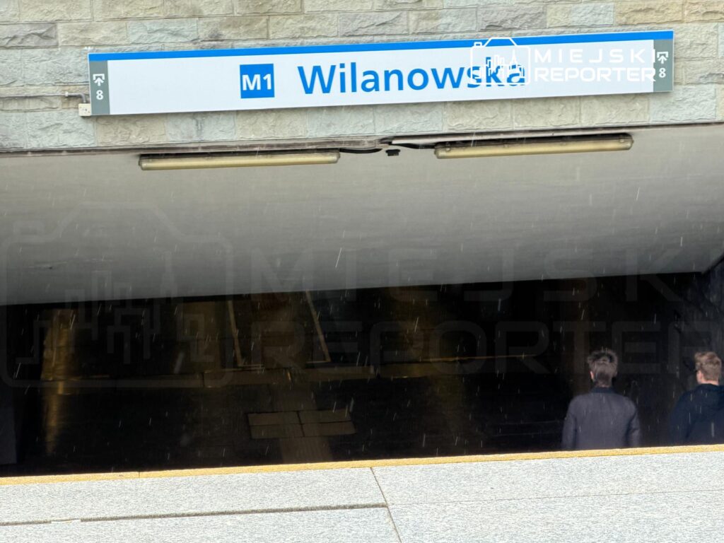 Dwaj mężczyźni w ciemnych ubraniach wchodzą do stacji metra pod znakiem "Wilanowska".