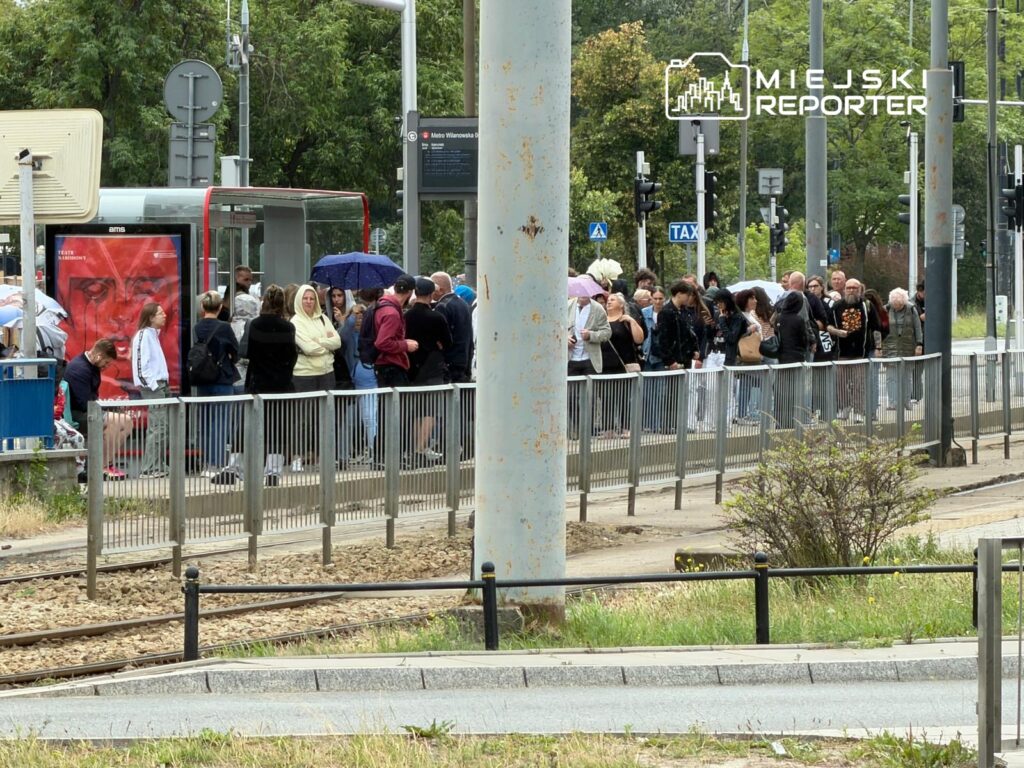 Grupa osób czeka w kolejce na przystanku tramwajowym, niektórzy trzymają parasole, w tle widoczny kiosk.