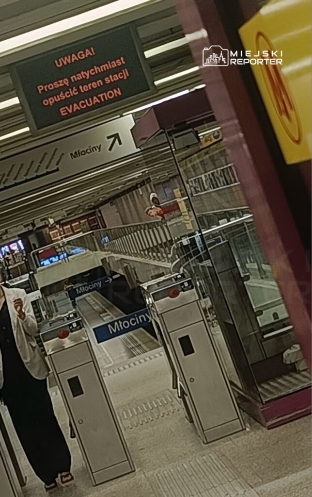 Na stacji metra widoczny jest znak ostrzegawczy z napisem "Uwaga! Proszę natychmiast opuścić teren stacji EVACUATION" oraz przejście do peronu.