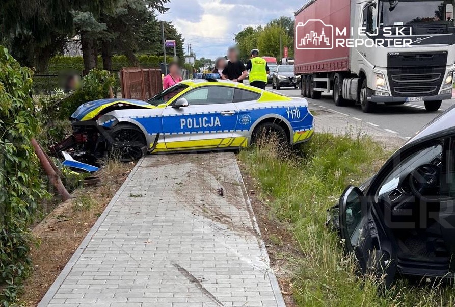 Policyjny samochód osobowy stoi na trawniku obok drogi, a funkcjonariusze rozmawiają z uczestnikami zdarzenia.