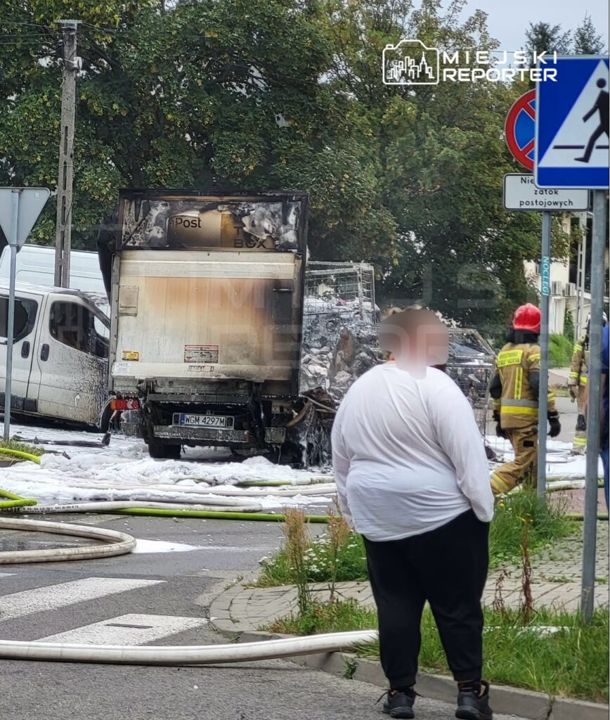 Przedstawiciel służb ratunkowych gasi pożar spalonego pojazdu dostawczego na ulicy, obok stoi osoba w białej koszuli.