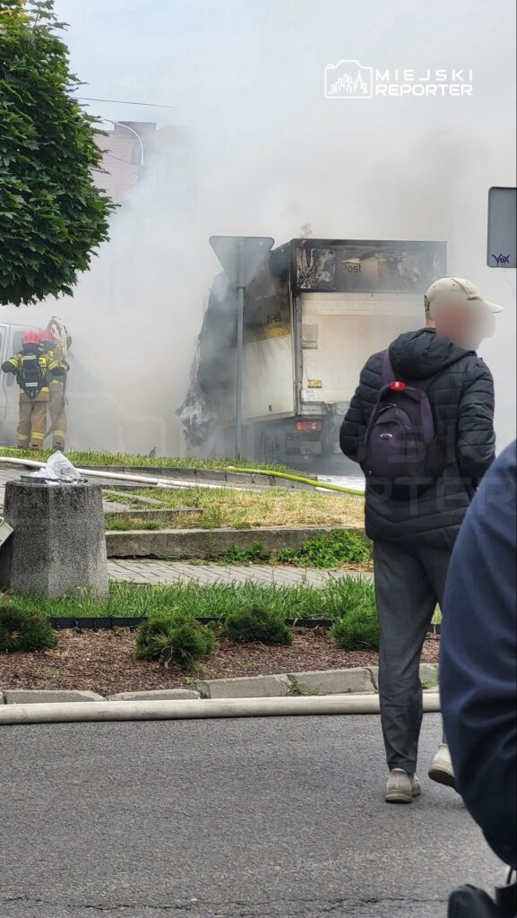 Strażacy gaszą palący się pojazd dostawczy, podczas gdy przechodzień obserwuje sytuację w otoczeniu zieleni.