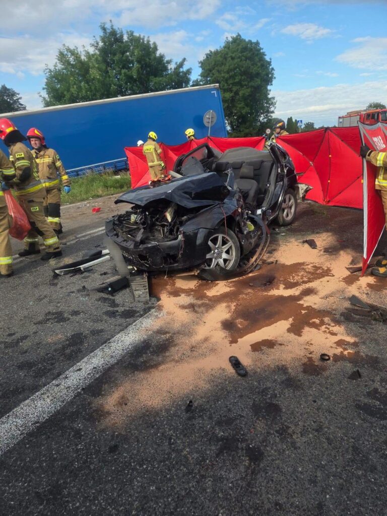 W miejscu wypadku drogowego strażacy pracują nad rozbitym samochodem osobowym, obok widoczny ciężarówka.