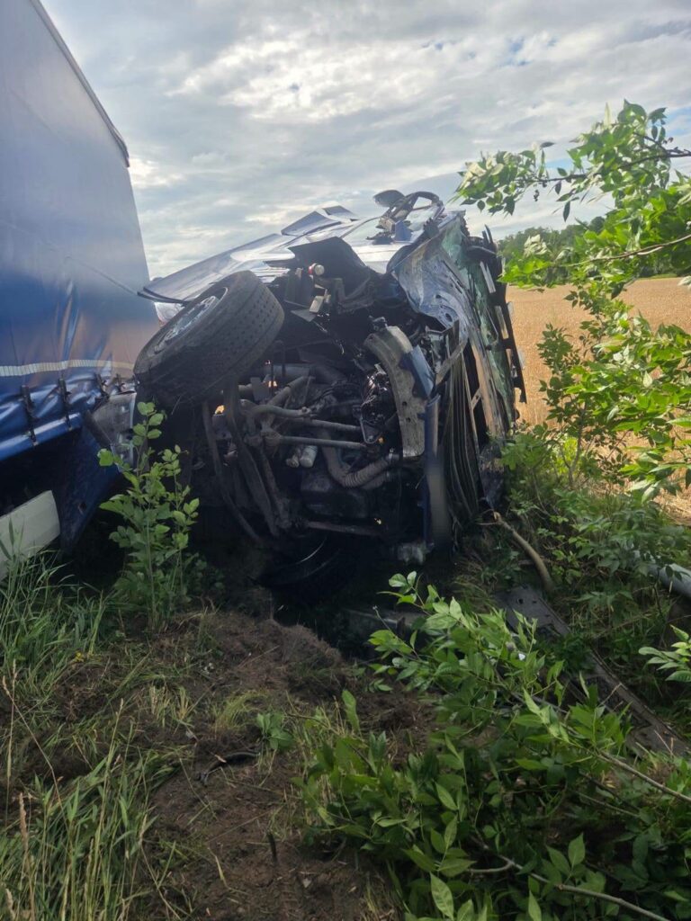 Nieoznakowana ciężarówka leży na boku w pobliżu drogi, otoczona zielenią i polami.