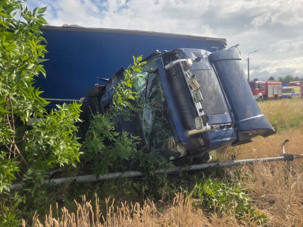 Tir przewrócony na bok w zaroślach przy drodze, z uszkodzoną kabiną i widocznymi służbami ratunkowymi w tle.