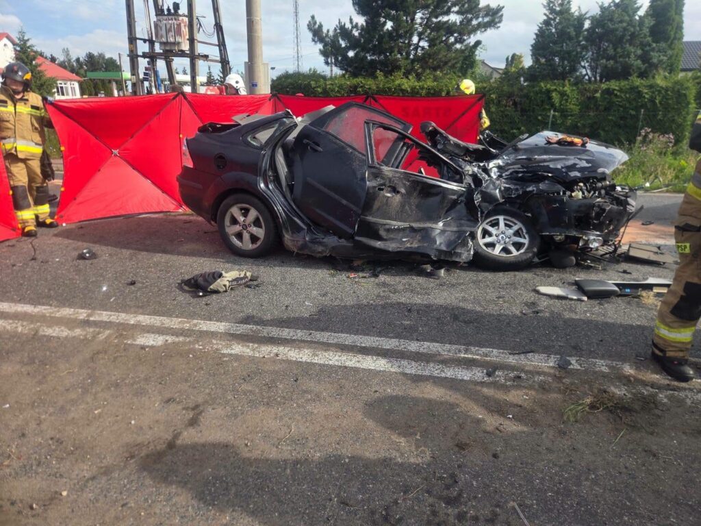 W pobliżu drogi znajduje się zniszczony samochód osobowy, otoczony przez strażaków i czerwone osłony.