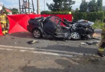 W pobliżu drogi znajduje się zniszczony samochód osobowy, otoczony przez strażaków i czerwone osłony.