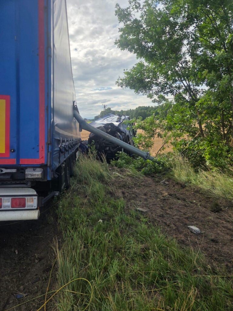 Czarny samochód osobowy w rowie, z uszkodzoną karoserią, obok niebieska ciężarówka stojąca na poboczu.