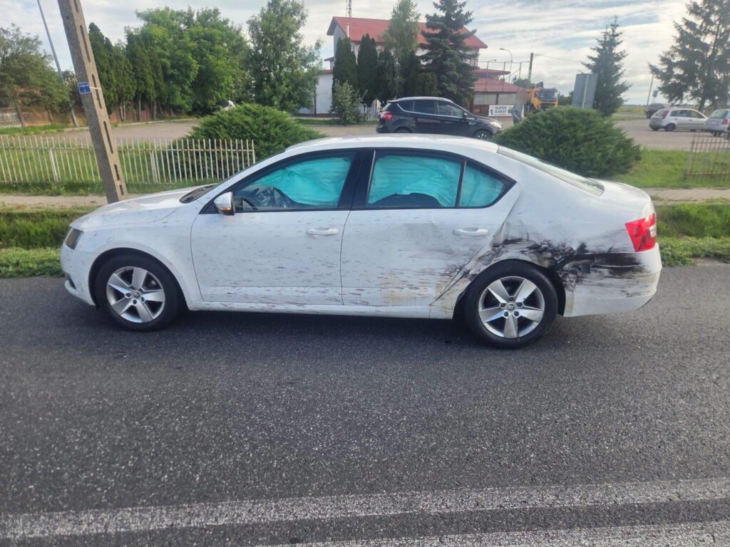 Biały samochód osobowy z uszkodzeniami na boku zaparkowany na poboczu drogi, w tle drzewa i inne pojazdy.