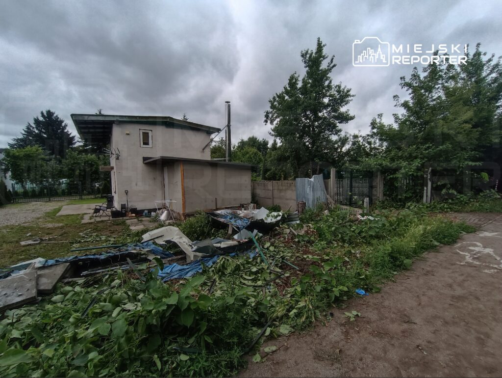 Zaniedbany ogród z porośniętymi chwastami i odpadami budowlanymi, obok nowoczesny dom z płaskim dachem.
