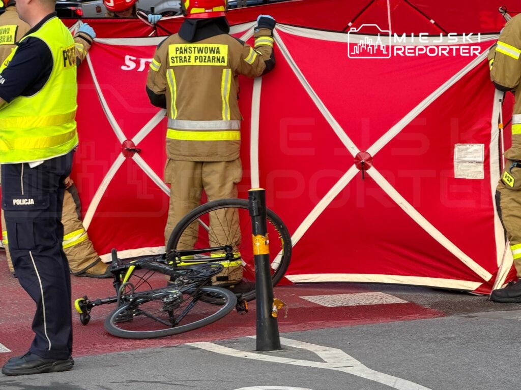Strażacy w mundurach ustawiają czerwony namiot na ulicy, obok leży uszkodzony rower.