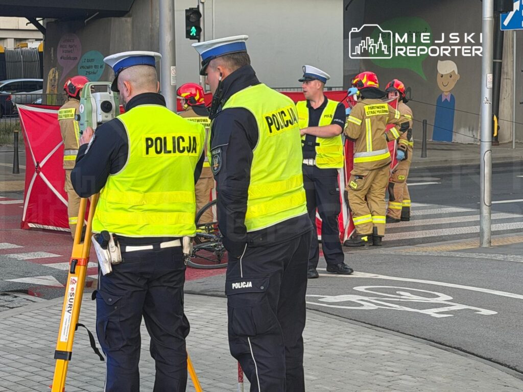 Policjanci w kamizelkach odblaskowych prowadzą działania na miejscu wypadku, obok pracują strażacy.