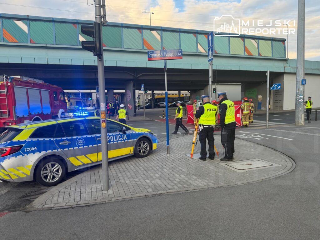 Grupa policjantów w kamizelkach odblaskowych zabezpiecza miejsce zdarzenia pod wiaduktem, obok wozu strażackiego.