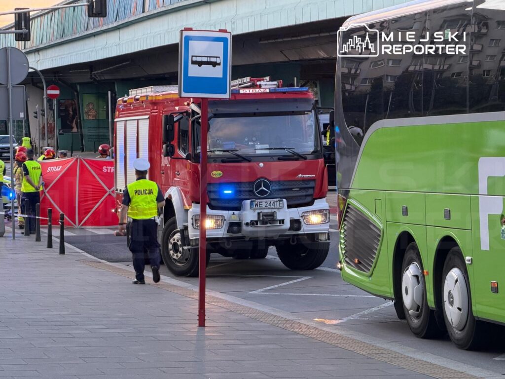 Strażacy w kaskach i mundurach pracują przy wozie strażackim, a policjant kieruje ruchem obok autobusu w mieście.