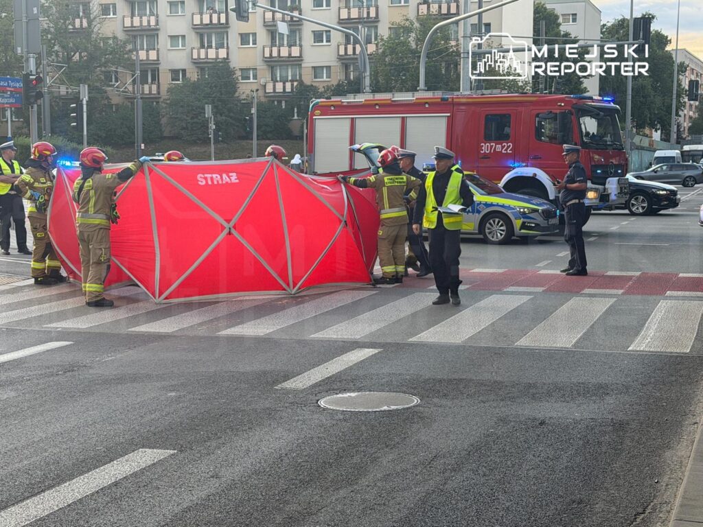 Grupa strażaków i policjantów ustawia czerwoną osłonę na pasach, podczas interwencji na ruchliwej ulicy.