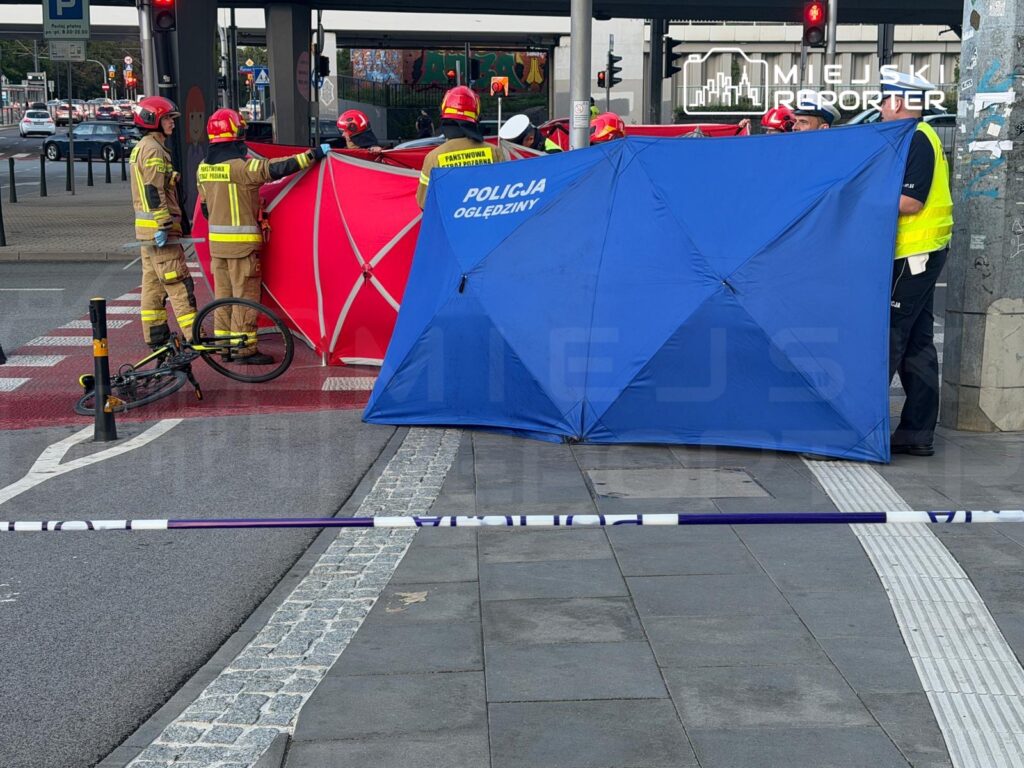 Grupa strażaków i policjantów ustawia czerwony i niebieski namiot na skrzyżowaniu, obok przewróconego roweru.