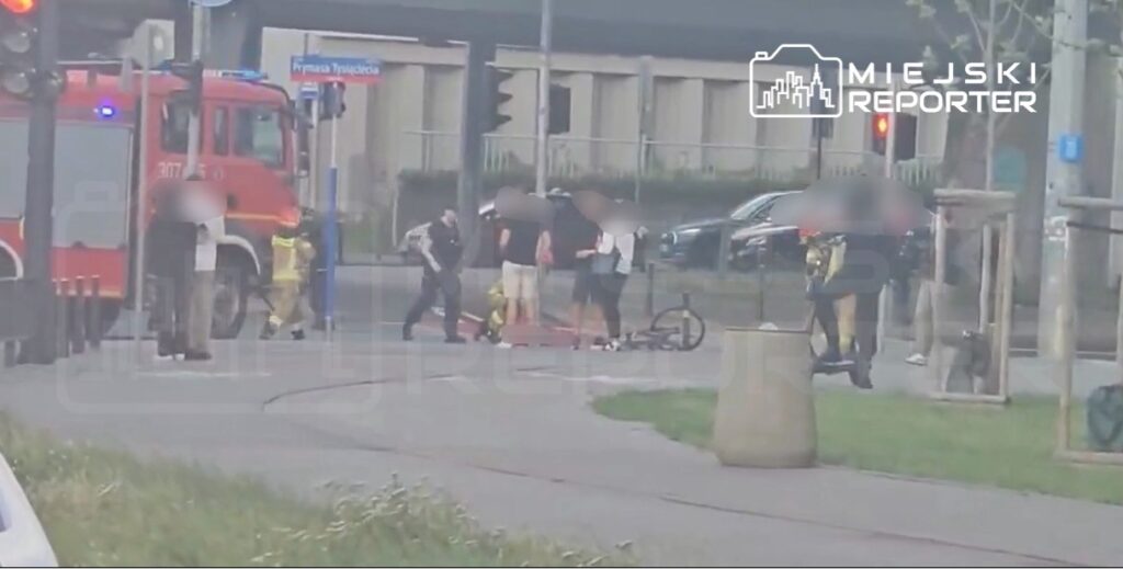 Grupa osób na chodniku rozmawia z funkcjonariuszami w pobliżu wozu strażackiego. Na ziemi leży rower.
