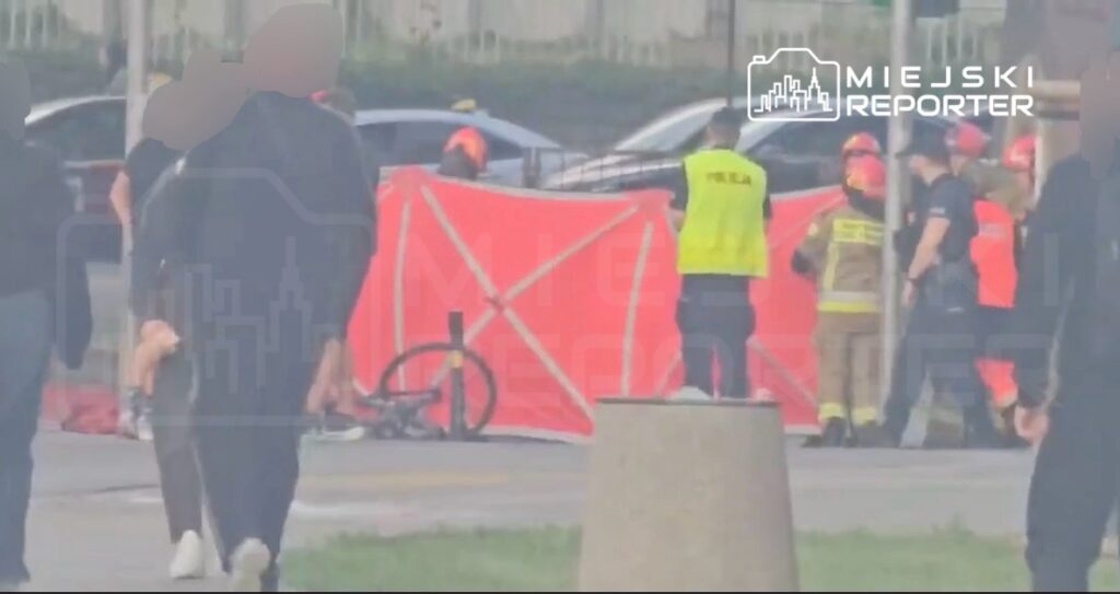 Grupa ratowników i policjantów pracuje przy wypadku drogowym, zasłoniętym czerwoną plandeką, obok leżącego roweru.