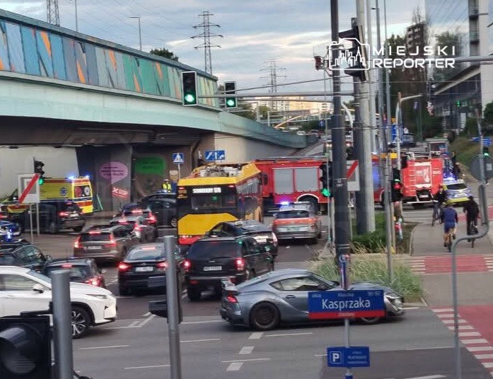 Karetki pogotowia i wóz strażacki stoją w zablokowanym ruchu drogowym pod wiaduktem, w tle widoczne sygnalizatory świetlne.