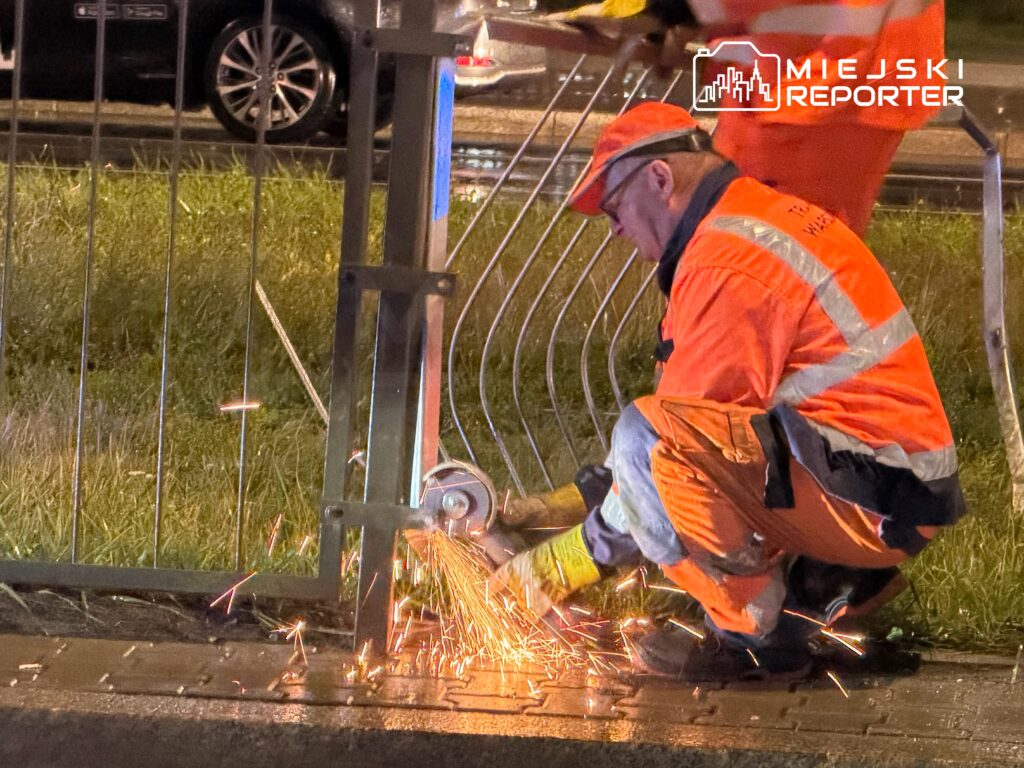 Pracownik w pomarańczowym uniformie ścina metalowy słupek, generując iskry, na ulicy w nocy.