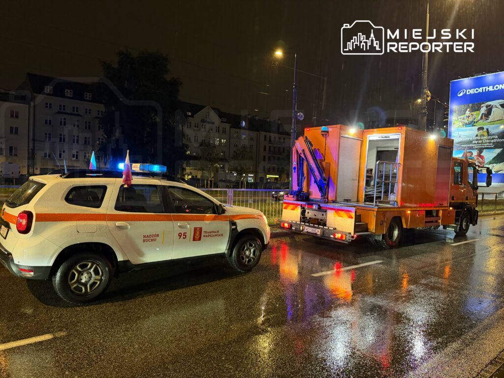 Samochód służbowy z niebieskim światłem zatrzymany na deszczowej ulicy obok pomarańczowego pojazdu technicznego.