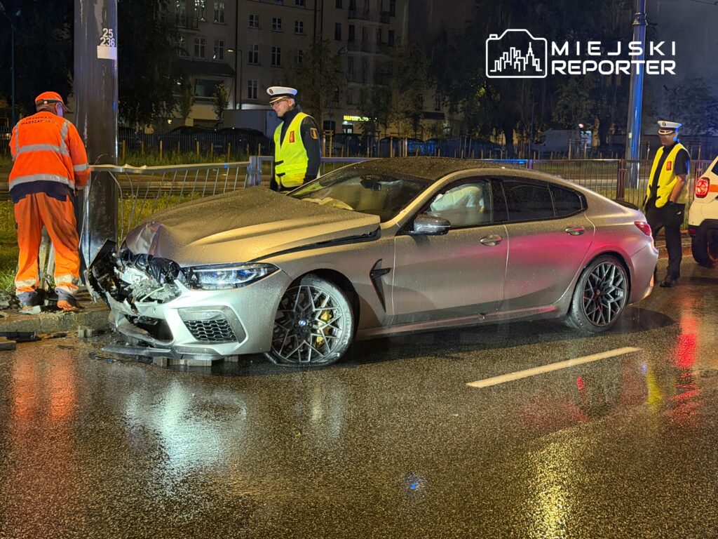 Uszkodzony samochód osobowy stoi przy latarni, a obok pracują strażacy i policjanci w odblaskowych kamizelkach.