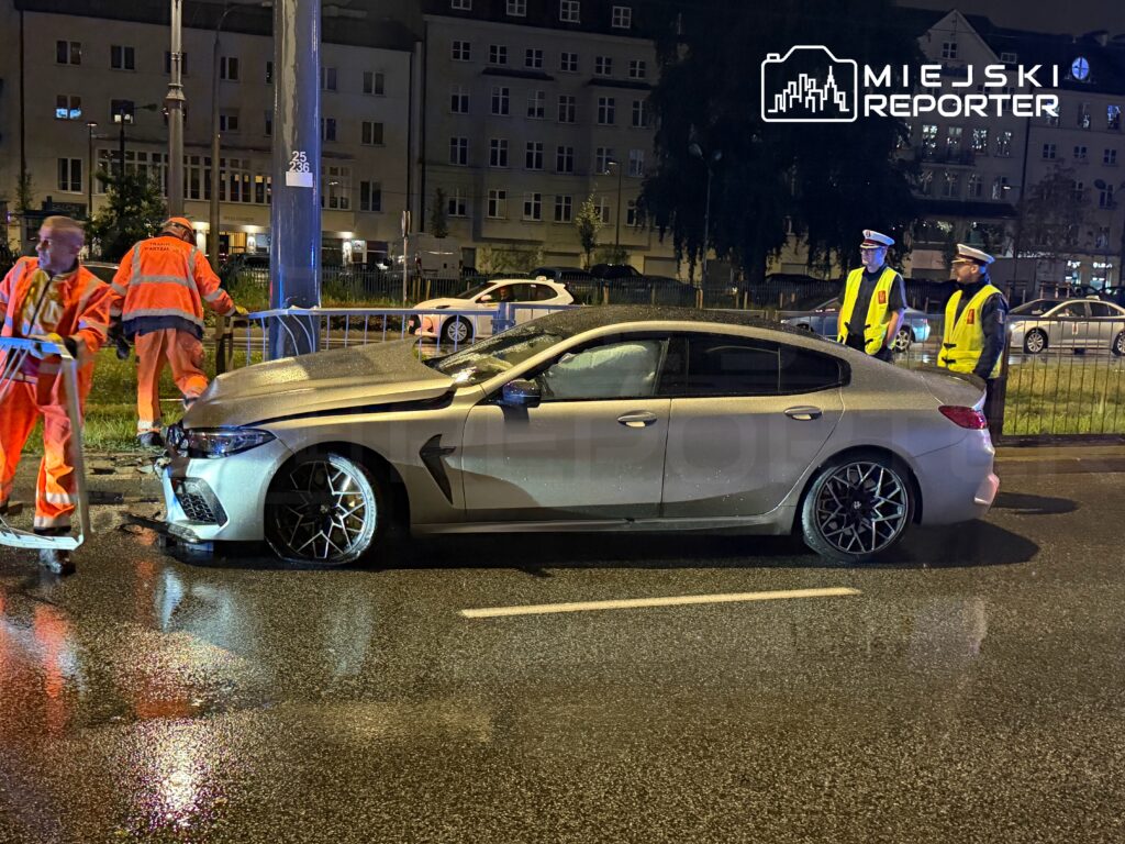 Grupa pracowników w odblaskowych kamizelkach zabezpiecza miejsce wypadku, gdzie uszkodzone auto stoi na mokrej jezdni.