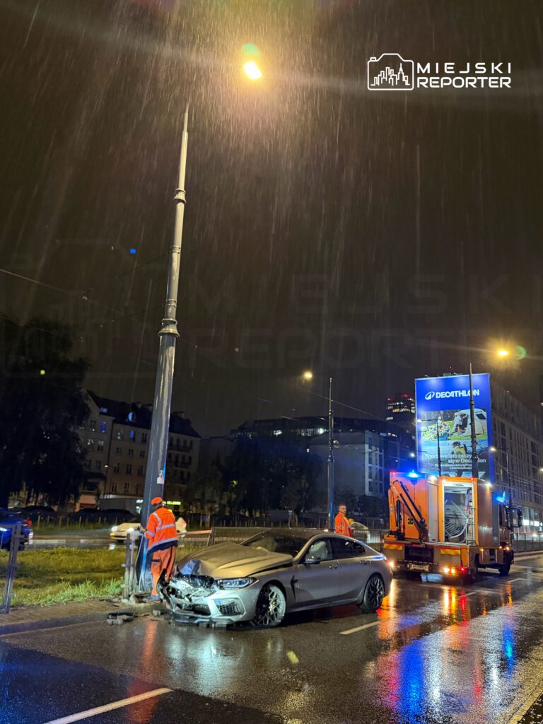 W nocy, w deszczu, strażacy pracują przy uszkodzonym samochodzie stojącym obok latarni ulicznej.