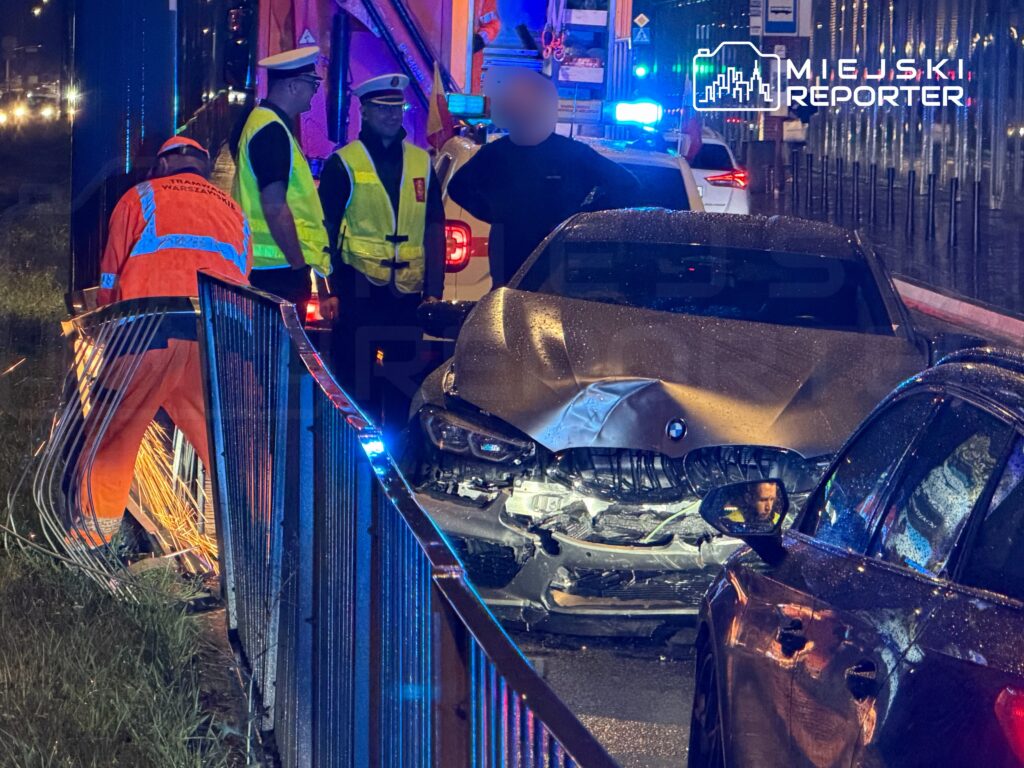 Mężczyzna w odzieży roboczej usuwa uszkodzenia bariery, obok stoi zespół policji przy wypadku drogowym z uszkodzonym samochodem.