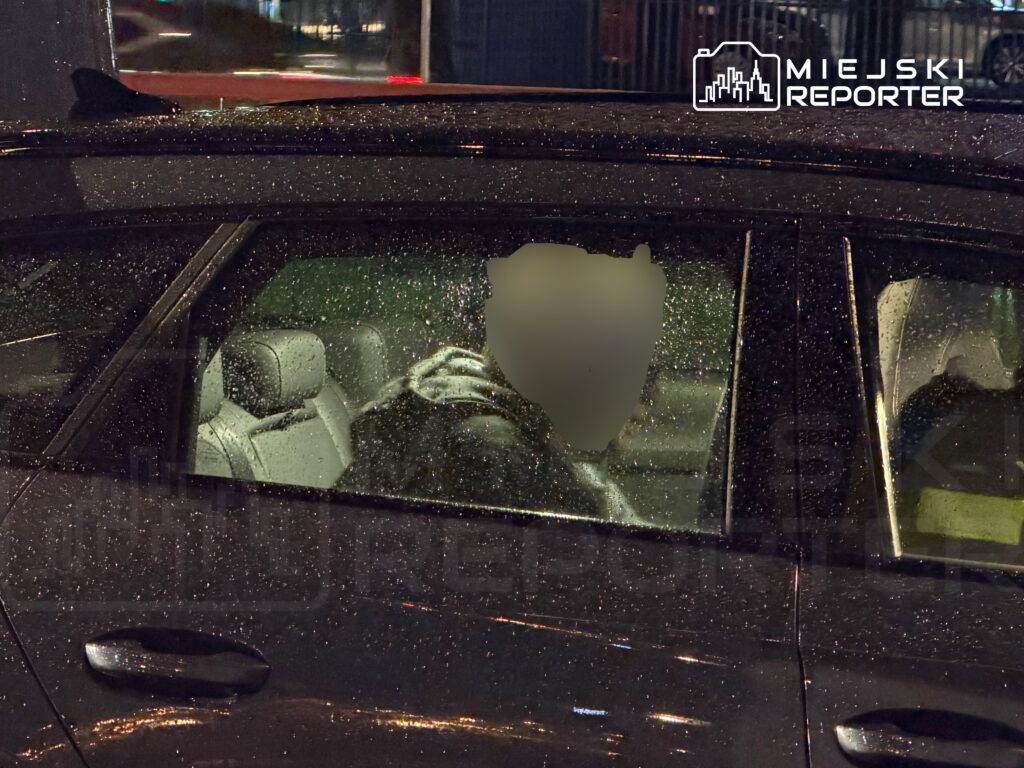 Kierowca w samochodzie z widocznymi kroplami deszczu na szybie, w nocy w miejskim otoczeniu.