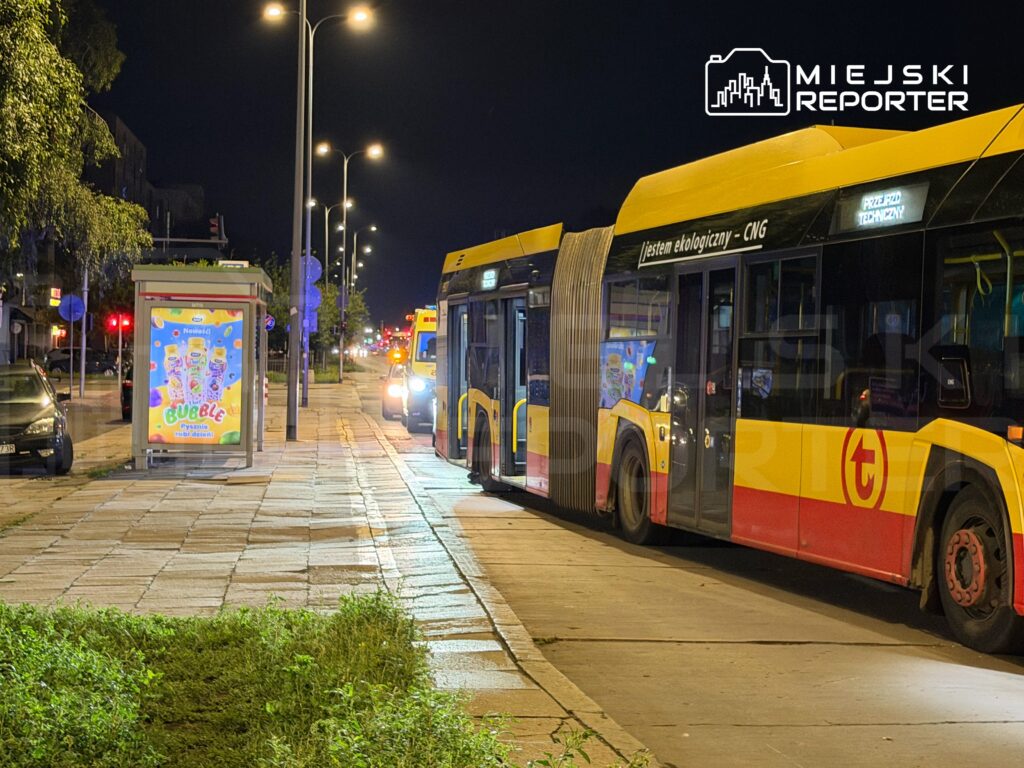 Dwa autobusy stoją na przystanku nocą, obok widać przeszkloną wiatę z kolorowym plakatem reklamowym.