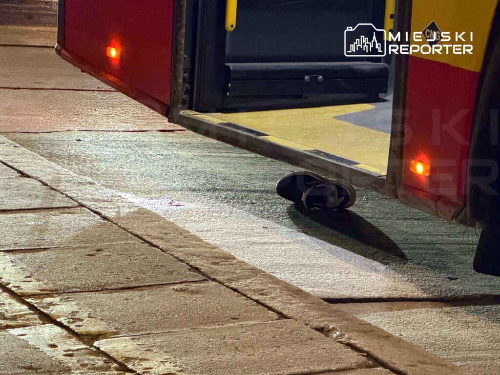 Otwarte drzwi autobusu stojącego na mokrej nawierzchni, z widoczną częścią podwozia i oświetleniem.
