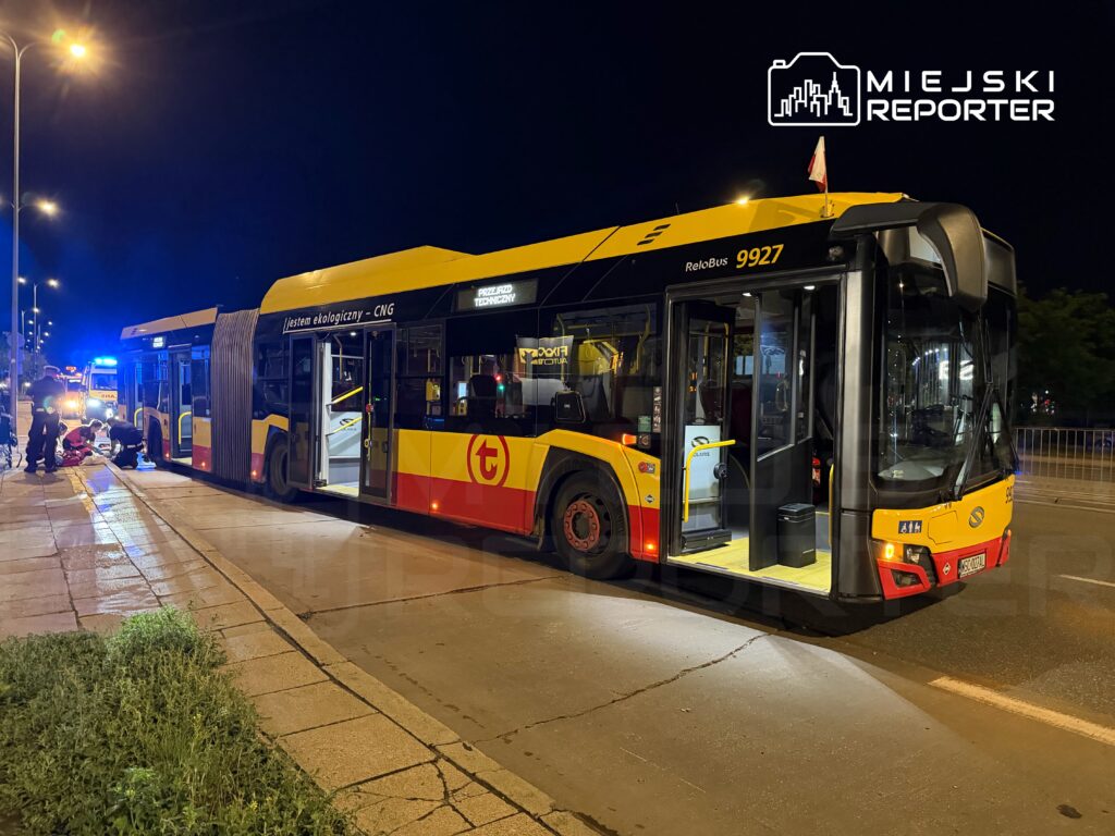 Autobus miejski stojący na ulicy w nocy, obok grupy osób pomagających poszkodowanemu. W tle widać radiowóz z włączonymi sygnałami świetlnymi.