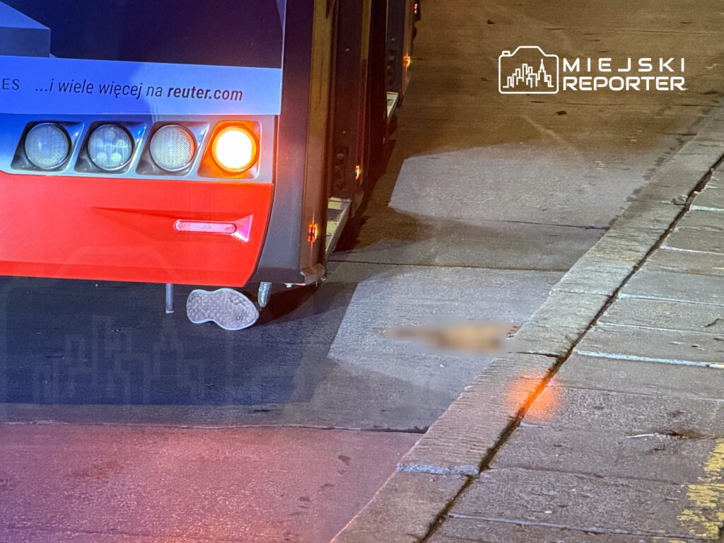 Autobus z czerwono-niebieską karoserią stoi na ulicy, z tyłu widoczna odblaskowa lampa. Na jezdni leży odciśnięty ślad buta.