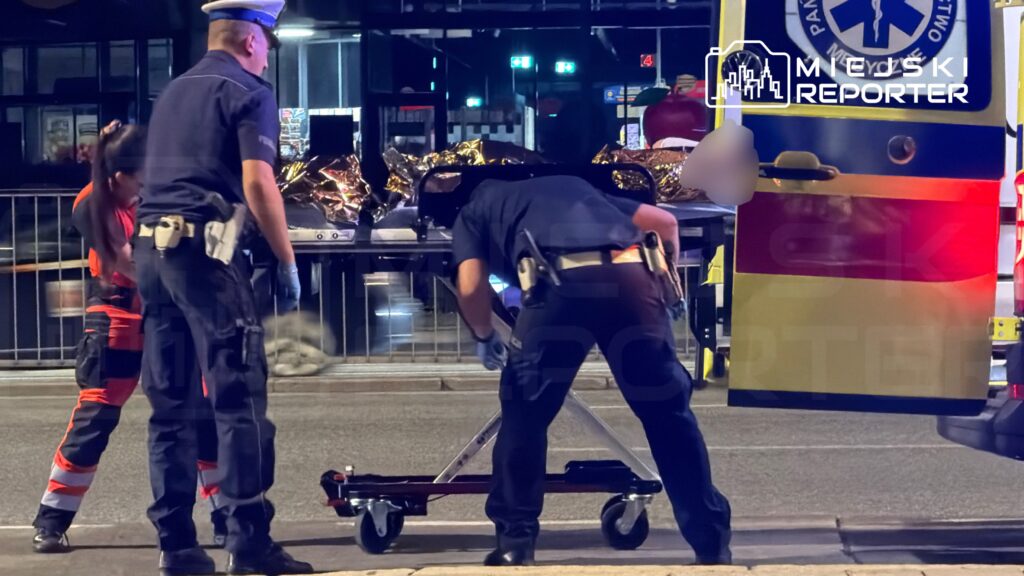 Ratownicy medyczni i policjanci transportują osobę na noszach do karetki na oświetlonej ulicy nocą.