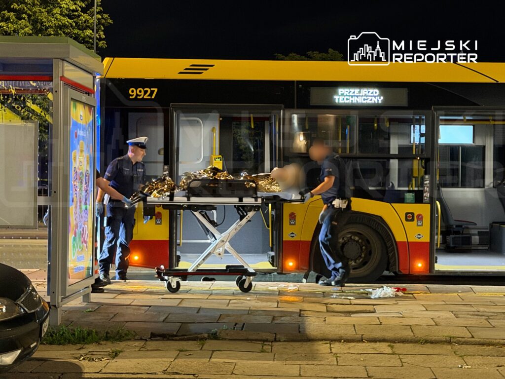 Dwaj mężczyźni w mundurach transportują osobę na noszach obok autobusu w nocy, z przystankiem w tle.