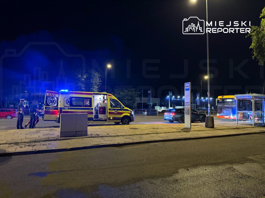 Ambulans z włączonymi sygnałami świetlnymi stoi na ulicy, obok dwóch policjantów rozmawiających przy otwartych drzwiach pojazdu.