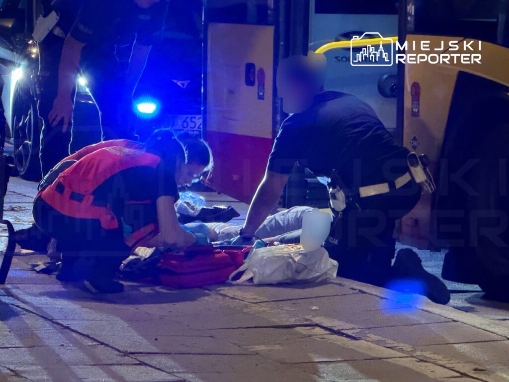 Ratownicy medyczni udzielają pomocy poszkodowanemu na ulicy, w otoczeniu pojazdów i niebieskich świateł sygnalizacyjnych.