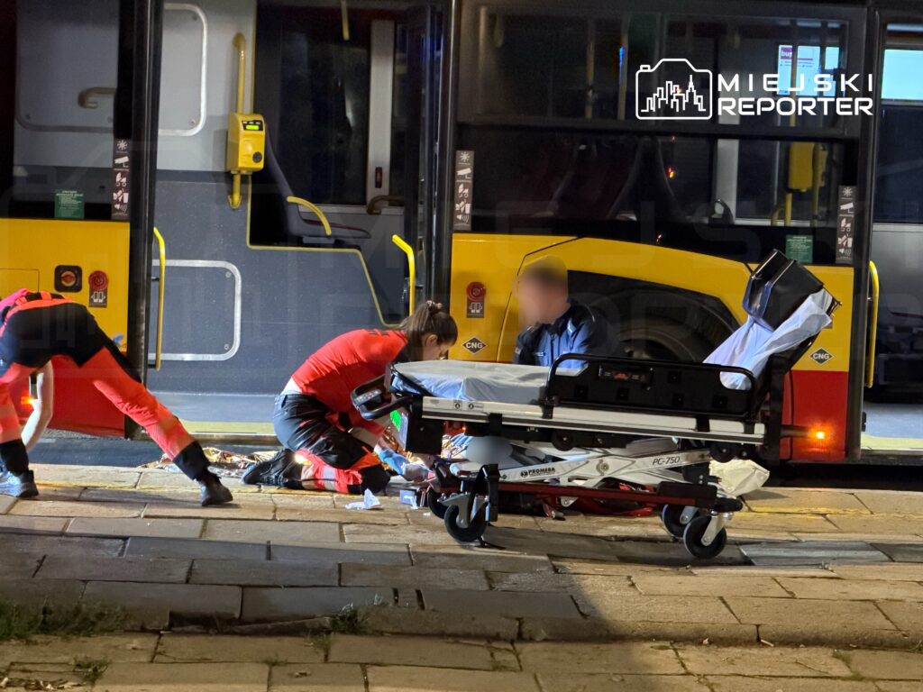Ratownicy medyczni udzielają pomocy osobie na chodniku obok autobusu miejskiego w nocy.