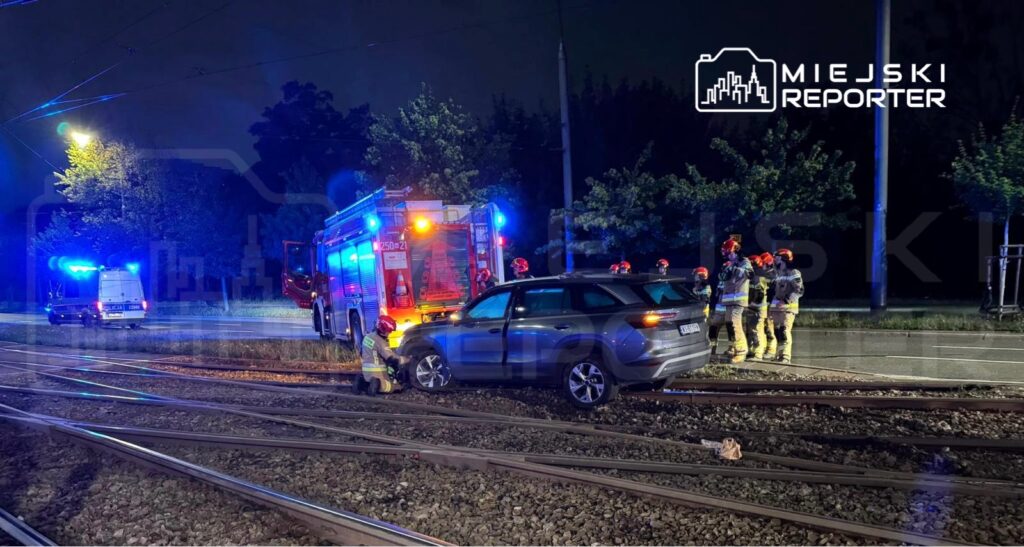 Strażacy w kaskach rozmawiają przy unieruchomionym samochodzie na torach tramwajowych w nocy.