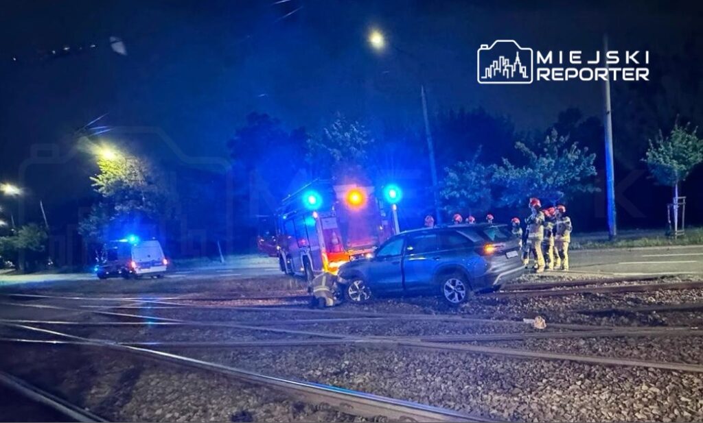 Na torach kolejowych stoi uszkodzony samochód, obok grupka strażaków i pojazd strażacki z włączonymi sygnałami świetlnymi.