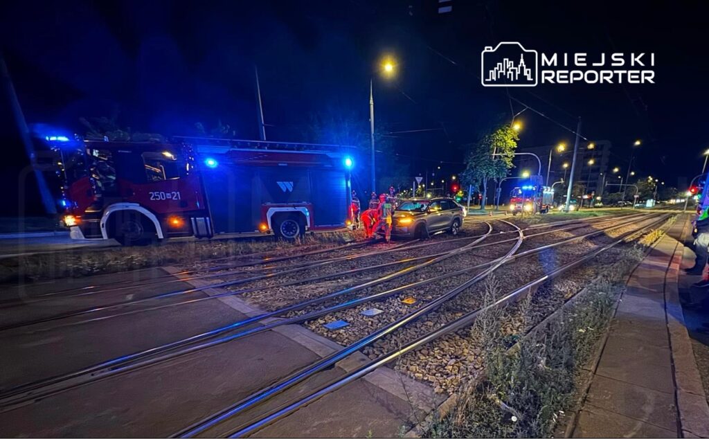 Strażacy w odblaskowych kamizelkach udzielają pomocy kierowcy w samochodzie na torach tramwajowych w nocy.