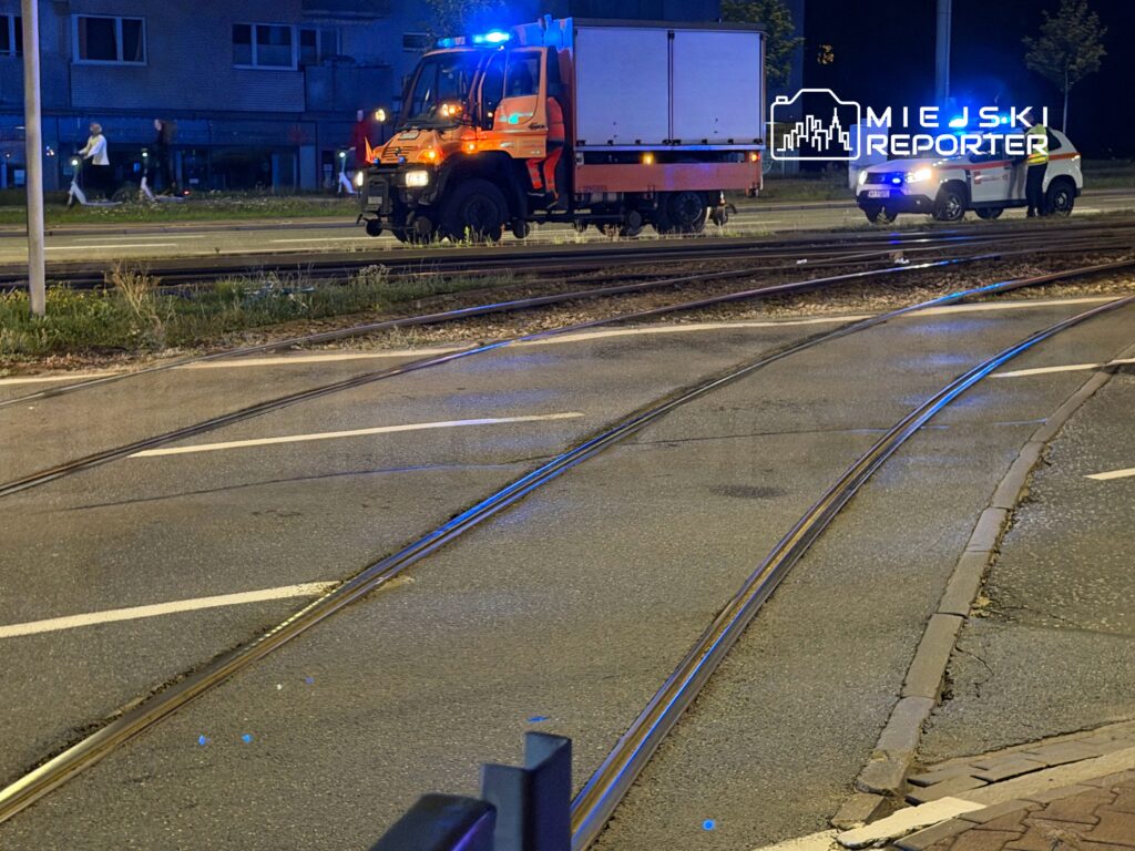 Pojazd ratunkowy z włączonymi sygnałami świetlnymi stoi obok torów tramwajowych w nocy. W tle przechodnie spacerują chodnikiem.