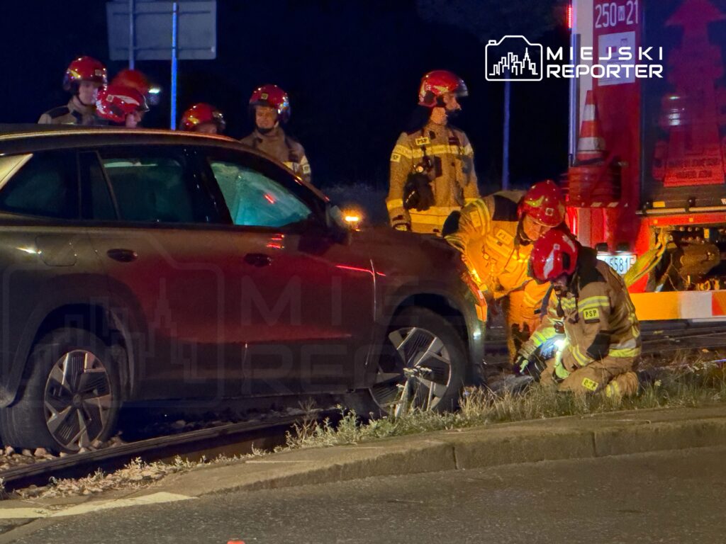Grupa strażaków w hełmach pomaga przy uszkodzonym samochodzie, który stoi na torach kolejowych w nocy.