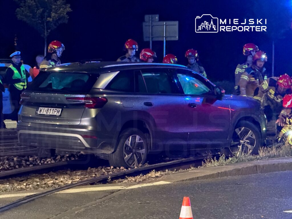 Samochód osobowy stoi na torach kolejowych, a wokół niego pracują strażacy w hełmach oraz policjanci.